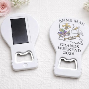 May include: Two white bottle openers with a magnetic back. One shows a graphic of the state of Louisiana, a fleur-de-lis, a mask, a pelican, and a trumpet, with the text ANNE MAE GRANDS WEEKEND 2026. The other shows the plain white back with a magnet.