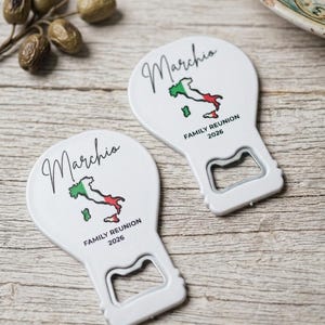 May include: Two white bottle openers with a map of Italy in green, white, and red. The word "Marchio" is written in cursive above the map. Below the map, the text reads "FAMILY REUNION 2026". The openers are on a wooden surface.