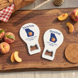 May include: Two white bottle openers with a paddle shape. Each features a blue outline of the state of Georgia, a peach graphic, and the text Newton FAMILY REUNION 2026 printed in dark blue. The openers have a rectangular cutout at the base.