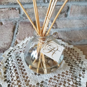 May include: A clear glass diffuser bottle filled with pebbles and reeds, with a tag that reads "Candles By Renee, Handmade, Harford, WI". The bottle sits on a cream-colored, open-weave doily, against a brick wall background.