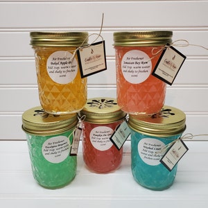 May include: Six glass jars with gold lids and decorative labels. Each jar contains a colorful gel-like substance and is labeled with a different scent: Baked Apple Pie, Jamaican Bay Rum, Eucalyptus Spearmint, Pumpkin Pie Spice, Washed Linen. The jars are arranged in a semi-circle on a white surface.