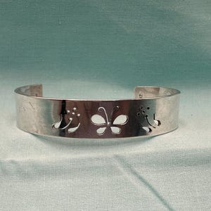 May include: A silver-toned metal cuff bracelet featuring a cutout design of a central butterfly flanked by two floral motifs. The band has a smooth, polished finish and an open back, showcasing a clean, metallic aesthetic with precise cutouts.
