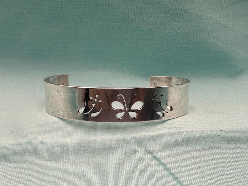 May include: A silver-toned metal cuff bracelet featuring a cutout design of a central butterfly flanked by two floral motifs. The band has a smooth, polished finish and an open back, showcasing a clean, metallic aesthetic with precise cutouts.