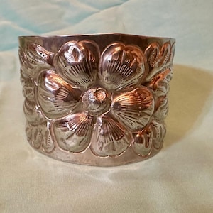 May include: Silver-colored metal cuff bracelet with a floral design. The bracelet features embossed flowers around the entire band. The cuff is a cylinder shape and sits on a light green surface.