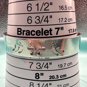 May include: A silver-toned metal cuff bracelet featuring a cut-out butterfly design flanked by floral motifs. The bracelet is positioned around a white sizing tool that displays measurements ranging from 6.5 inches to 8.25 inches for reference.