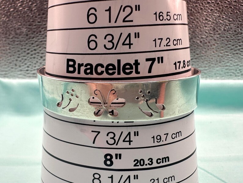 May include: A silver-toned metal cuff bracelet featuring a cut-out butterfly design flanked by floral motifs. The bracelet is positioned around a white sizing tool that displays measurements ranging from 6.5 inches to 8.25 inches for reference.