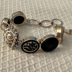 May include: A silver-toned link bracelet featuring a series of circular and square charms. The charms include black discs with embossed floral patterns, a square charm encrusted with clear rhinestones in a spiral design, and a toggle clasp closure.