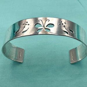 May include: A silver cuff bracelet featuring a cutout butterfly design in the center flanked by two cutout floral motifs. The interior of the band is stamped with the word STERLING and the maker mark CHAVEZ on the ends of the open cuff.