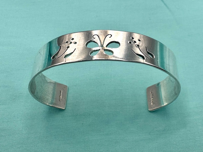 May include: A silver cuff bracelet featuring a cutout butterfly design in the center flanked by two cutout floral motifs. The interior of the band is stamped with the word STERLING and the maker mark CHAVEZ on the ends of the open cuff.