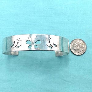 May include: A silver-colored metal cuff bracelet featuring a cutout design of a central butterfly flanked by two floral motifs. A United States dime is positioned to the right of the bracelet to provide a visual scale for the item's size.