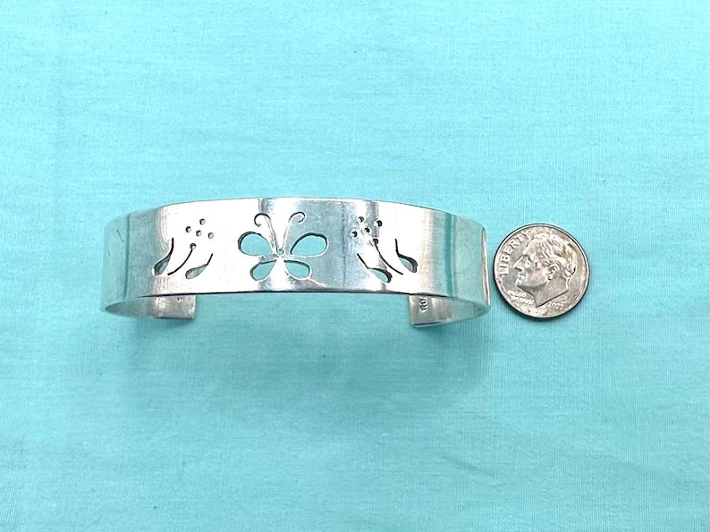 May include: A silver-colored metal cuff bracelet featuring a cutout design of a central butterfly flanked by two floral motifs. A United States dime is positioned to the right of the bracelet to provide a visual scale for the item's size.