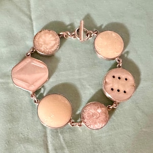 May include: Silver-toned bracelet featuring a series of light-colored, circular and geometric-shaped stones. The bracelet has a toggle clasp and is displayed on a light green surface. The stones have a textured appearance.