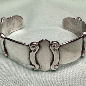 May include: A silver bracelet made from a repurposed spoon. The bracelet has a decorative design on the front and is open on the back.