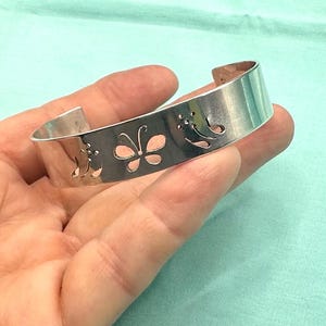 May include: A silver-toned cuff bracelet featuring a polished metallic finish and a series of cut-out designs, including a central butterfly motif flanked by stylized floral patterns, with an open back for adjustable wear.