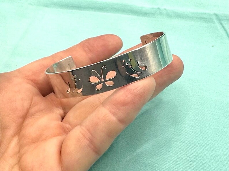 May include: A silver-toned cuff bracelet featuring a polished metallic finish and a series of cut-out designs, including a central butterfly motif flanked by stylized floral patterns, with an open back for adjustable wear.