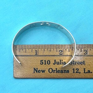May include: A silver cuff bracelet with a cutout floral pattern positioned over a wooden ruler. The bracelet measures approximately 2.5 inches in diameter across the opening, as shown by the markings on the ruler placed directly underneath it.