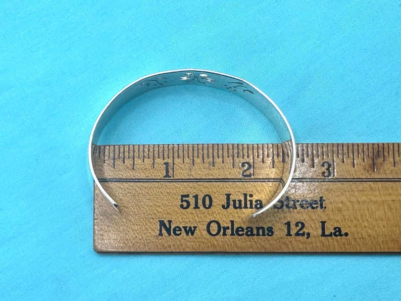 May include: A silver cuff bracelet with a cutout floral pattern positioned over a wooden ruler. The bracelet measures approximately 2.5 inches in diameter across the opening, as shown by the markings on the ruler placed directly underneath it.