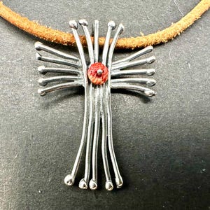 May include: A silver pendant necklace with a red and white center piece. The pendant is shaped like a cross with multiple thin metal strands extending from the center. The necklace is made of brown leather.
