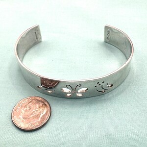 May include: A silver-colored metal cuff bracelet featuring cut-out butterfly designs along the band. The bracelet is positioned next to a United States dime coin for scale, showing the relative size of the jewelry piece against the small currency.
