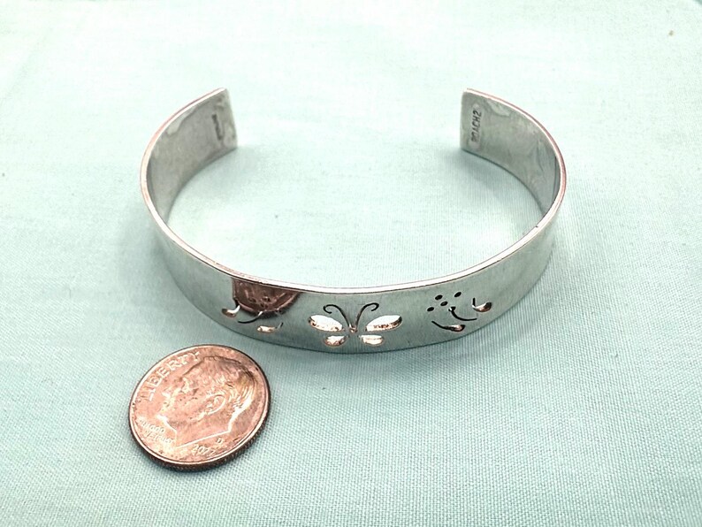 May include: A silver-colored metal cuff bracelet featuring cut-out butterfly designs along the band. The bracelet is positioned next to a United States dime coin for scale, showing the relative size of the jewelry piece against the small currency.