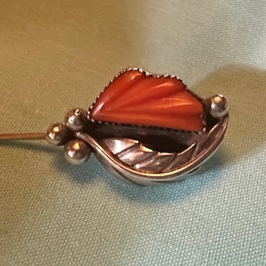 May include: A silver-toned hat pin with an orange, fan-shaped stone set in a decorative frame. The pin features a leaf design and small silver beads. The pin is on a light green background.