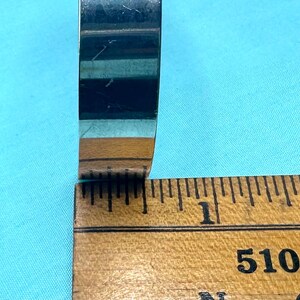 May include: A polished silver-toned metal ring band with a smooth, reflective surface. The ring is positioned against a wooden ruler, showing a width of approximately 0.25 inches. The band has a simple, flat profile with no visible engravings or stones.