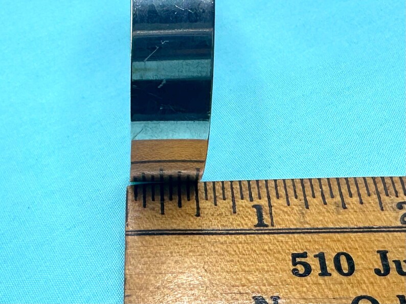 May include: A polished silver-toned metal ring band with a smooth, reflective surface. The ring is positioned against a wooden ruler, showing a width of approximately 0.25 inches. The band has a simple, flat profile with no visible engravings or stones.