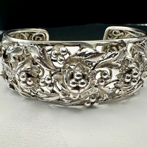 May include: A silver cuff bracelet with an open design. The bracelet is adorned with detailed floral and grape cluster motifs. The metal has a bright, reflective finish, and the bracelet appears to be unworn.
