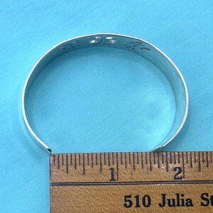May include: A silver-toned metal cuff bracelet featuring a series of cut-out floral or leaf-like shapes along the top edge. The bracelet is positioned against a wooden ruler, showing a diameter of approximately 2.5 inches across the opening.