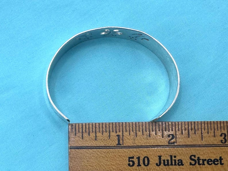 May include: A silver-toned metal cuff bracelet featuring a series of cut-out floral or leaf-like shapes along the top edge. The bracelet is positioned against a wooden ruler, showing a diameter of approximately 2.5 inches across the opening.