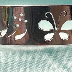 May include: A polished silver-tone metal cuff bracelet featuring a cutout design of a butterfly and a floral sprig with three small circular dots. The surface of the metal is reflective and smooth, showcasing the intricate die-cut shapes clearly.