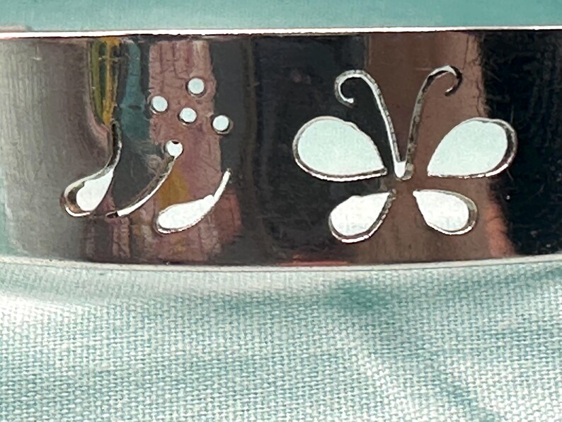May include: A polished silver-tone metal cuff bracelet featuring a cutout design of a butterfly and a floral sprig with three small circular dots. The surface of the metal is reflective and smooth, showcasing the intricate die-cut shapes clearly.