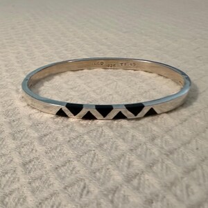 May include: A silver bangle bracelet featuring a repeating black triangular inlay pattern. The interior of the band is engraved with the text MEXICO 925 TF-49. The bracelet has a smooth, polished metallic finish and a circular shape.
