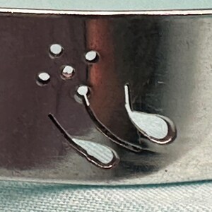 May include: A silver-toned metal cuff bracelet featuring a laser-cut design of a dandelion flower with delicate, curved stems and small circular cutouts representing seeds blowing in the wind, set against a smooth, reflective metallic surface.