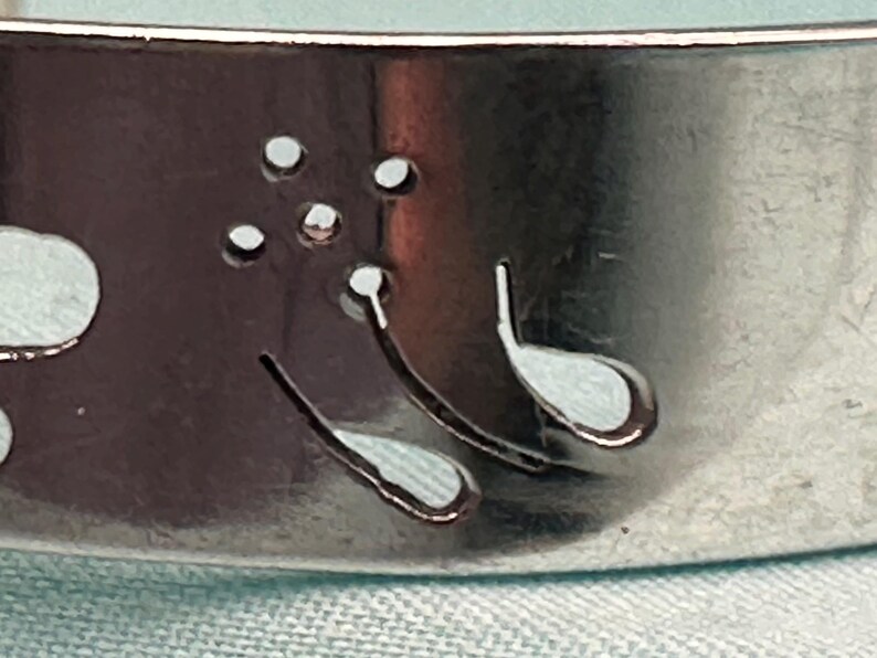 May include: A silver-toned metal cuff bracelet featuring a laser-cut design of a dandelion flower with delicate, curved stems and small circular cutouts representing seeds blowing in the wind, set against a smooth, reflective metallic surface.