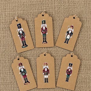 Peut inclure: Six étiquettes cadeaux rectangulaires en papier kraft brun, chacune ornée d'une illustration différente de casse-noisette. Les casse-noisettes portent des uniformes rouges, verts et dorés, avec des chapeaux hauts et des bâtons.