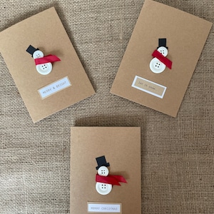 Könnte beinhalten: Drei handgefertigte Weihnachtskarten mit einem Schneemann-Design. Jede Karte besteht aus braunem Karton und zeigt einen Schneemann aus weißen Knöpfen, einem schwarzen Zylinder und einem roten Band-Schal. Die Karten haben den Text "Merry & Bright", "let it snow..." und "Merry Christmas".