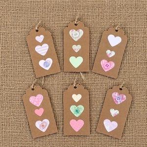 May include: Six rectangular brown kraft paper gift tags, each featuring three heart-shaped cutouts or appliqués arranged vertically. The hearts display various pastel colours, floral patterns, and stripes, with some adorned by small plastic buttons.