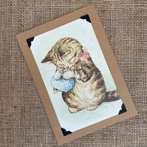 May include: A vintage postcard featuring a brown and white tabby cat holding a blue and white toy mouse. The cat is wearing a pink bow.