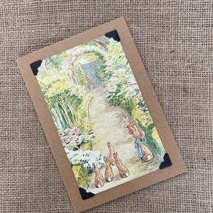 Könnte beinhalten: Eine Vintage-Postkarte mit einer farbenfrohen Illustration einer Gruppe von Kaninchen, die einen Gartenweg entlanglaufen. Die Kaninchen sind braun und weiß mit blauen Akzenten. Der Garten ist üppig grün mit rosa Blumen.