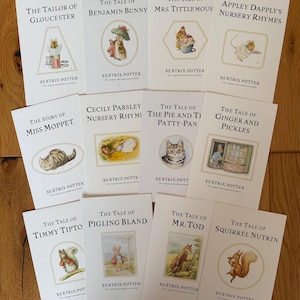May include: A collection of Beatrix Potter children's books, featuring titles like "The Tale of Peter Rabbit" and "The Tale of Benjamin Bunny." Each book has a white cover with illustrations and text. The books are arranged on a wooden surface.