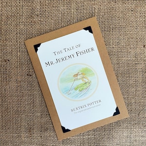 Könnte beinhalten: Ein brauner Papierumschlag mit einer weißen Karte darin. Die Karte zeigt eine Illustration eines Frosches auf einem Seerosenblatt mit dem Text "The Tale of Mr. Jeremy Fisher" und "Beatrix Potter The original and authorized edition".