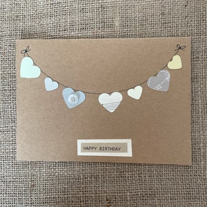 Könnte beinhalten: Eine handgemachte Karte mit braunem Hintergrund, auf der eine Girlande aus Papierherzen mit einem Knopf auf einem der Herzen zu sehen ist. Die Karte trägt den Schriftzug "Happy Birthday" in einem weißen Rechteck.