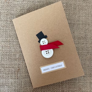 Könnte beinhalten: Eine handgefertigte Weihnachtskarte mit einem Schneemann-Design. Der Schneemann besteht aus weißen Knöpfen, einem roten Schleifenschal und einem schwarzen Zylinder. Die Karte ist naturfarben und trägt den Aufdruck "MERRY CHRISTMAS".