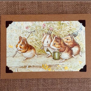 May include: A brown card with a watercolor illustration of four guinea pigs gardening. One guinea pig holds a rake, another a watering can, and a third a trowel. The card is framed with brown paper and black corner mounts.