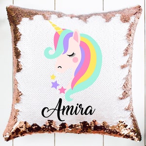 Könnte beinhalten: Ein weißes Kissen mit roségoldenen Pailletten, auf dem eine bunte Einhorn-Illustration und der Name "Amira" aufgedruckt sind.