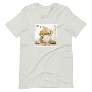 Puede incluir: Una camiseta blanca con un gráfico de un cachorro golden retriever bebiendo de una taza de café blanca. El texto "BUT FIRST COFFEE" está impreso en el gráfico.