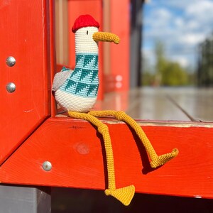 May include: A handmade crochet seagull doll with a red hat, teal and white patterned sweater, and yellow legs. The seagull is sitting on a red wooden surface, with a blurred background of a blue sky and trees.