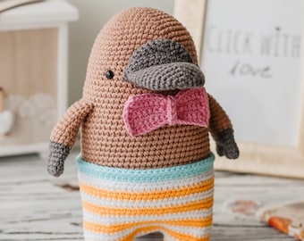 May include: A crocheted plush toy platypus wearing a pink bow tie and striped trousers. The toy is brown, grey, and blue.