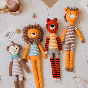 May include: Four crocheted stuffed animals: a lion, a tiger, a giraffe, and a monkey. The lion is orange with a blue and white striped shirt. The tiger is orange and black with a white striped shirt. The giraffe is brown and white with a white striped shirt. The monkey is brown and white with a white striped shirt. The animals are all lying on a white wooden surface.